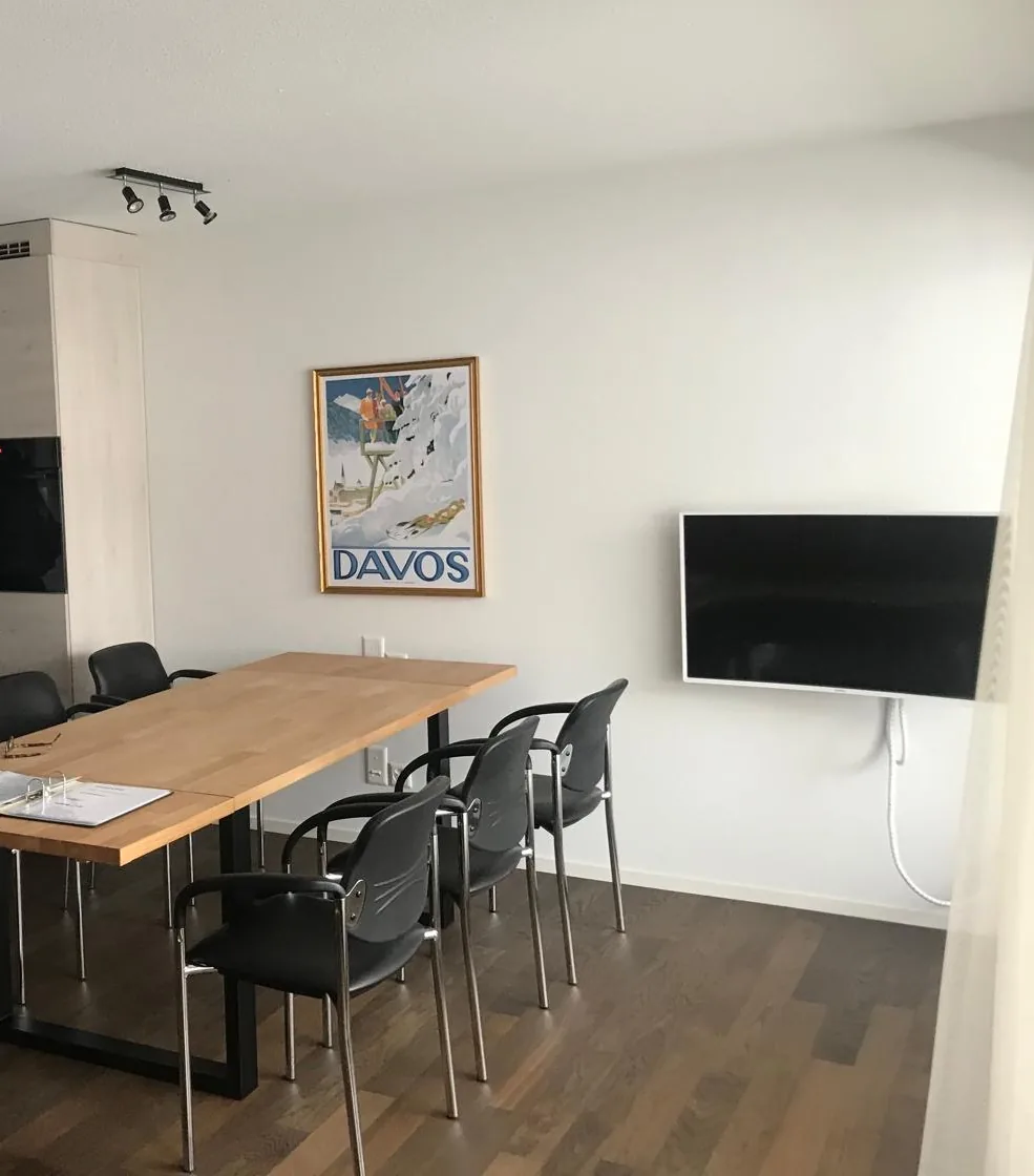 A meeting table with black conference chairs, a wall-mounted screen and a vintage Davos poster in an apartment living room