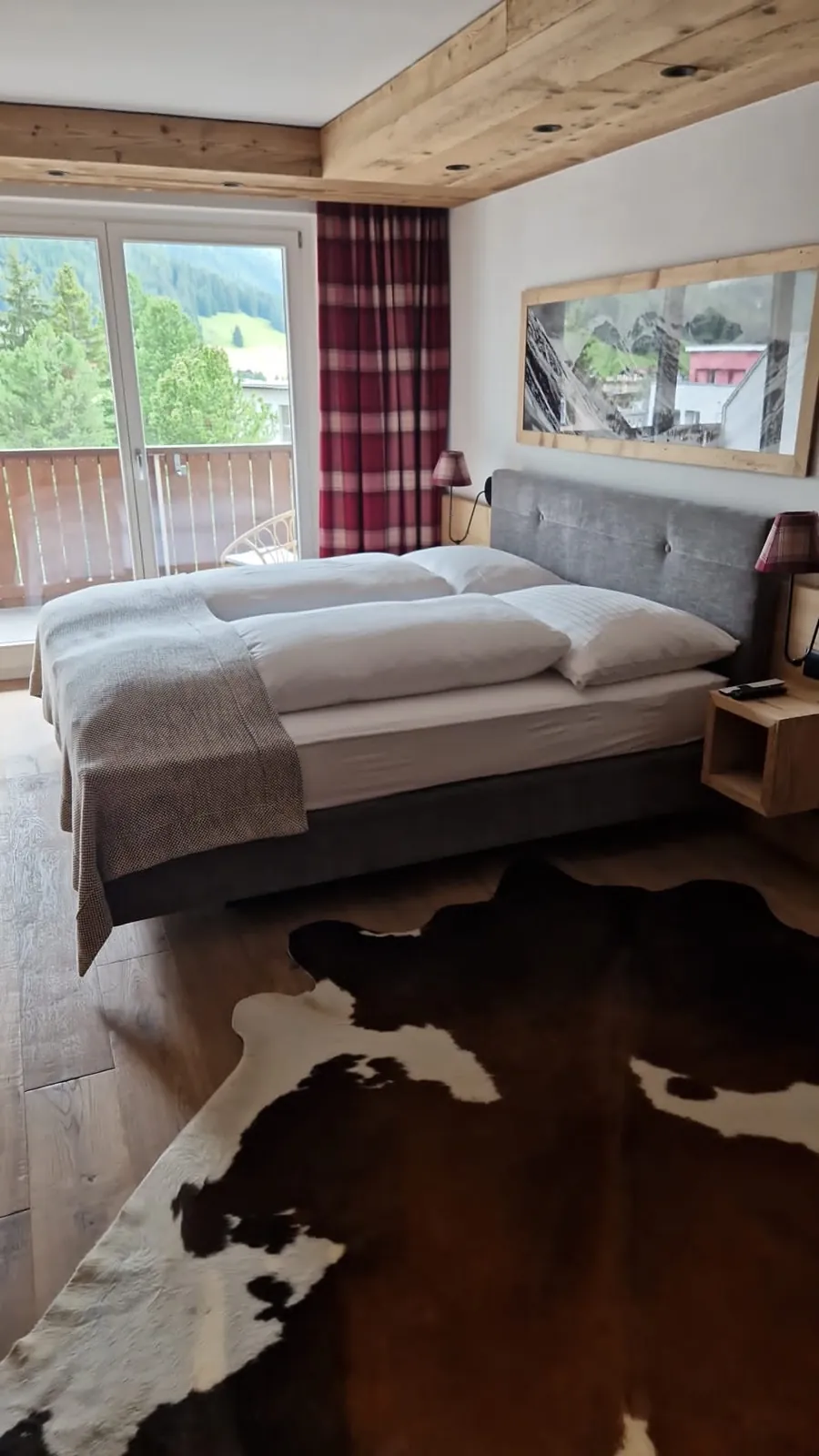 Double bedroom with a timber ceiling, tartan curtains, a cowhide rug and a balcony door onto pine trees and meadow