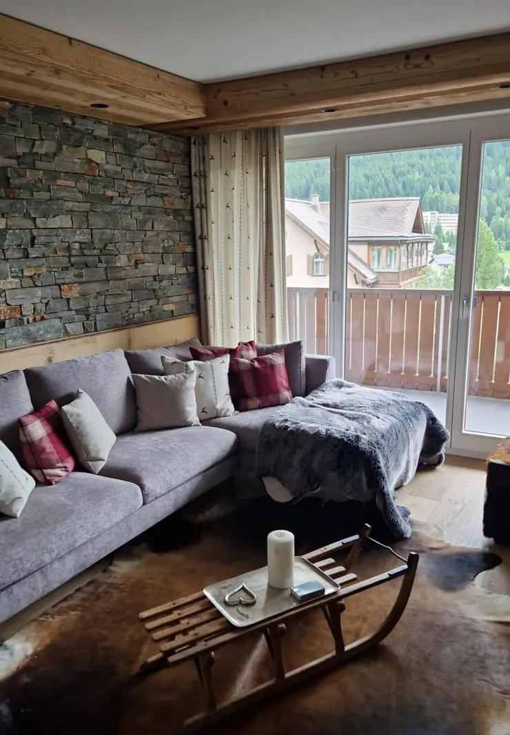 Chalet living room with a stone wall, a grey corner sofa with tartan cushions, a sledge used as a coffee table and a balcony door onto pine forest