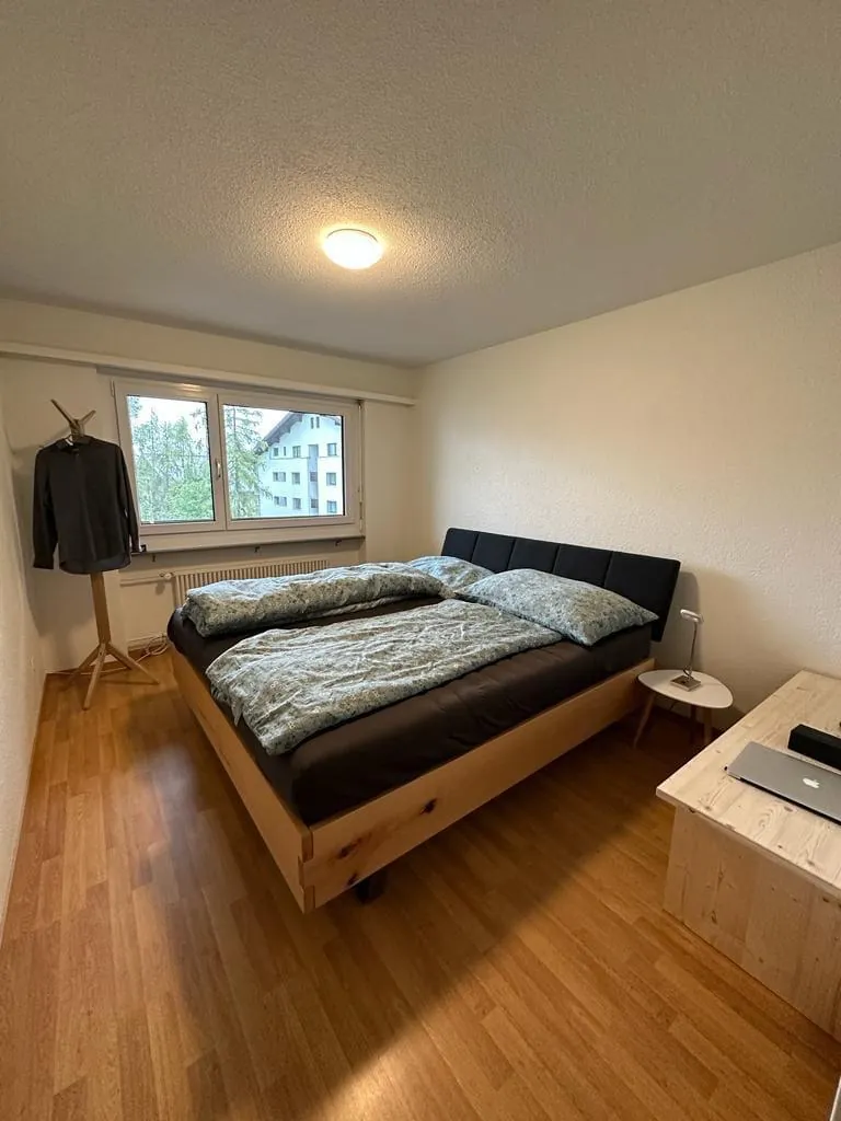 A double bedroom with a wooden bed, a desk and a window onto pine trees in a Davos apartment