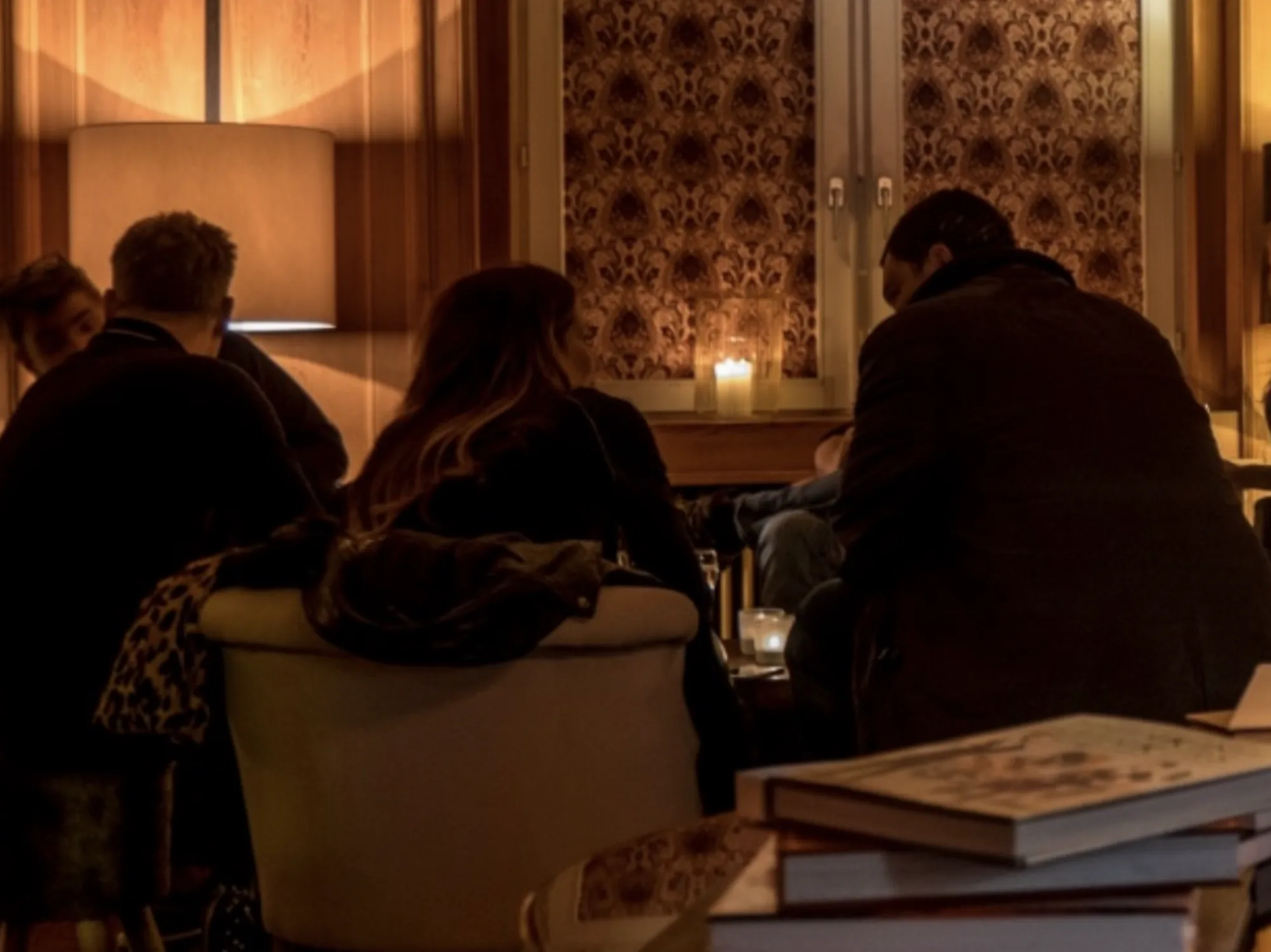 A small group seated in low armchairs by candlelight, wood panelling and a lit fireplace mantel, deep in conversation at a Davos fireside chat