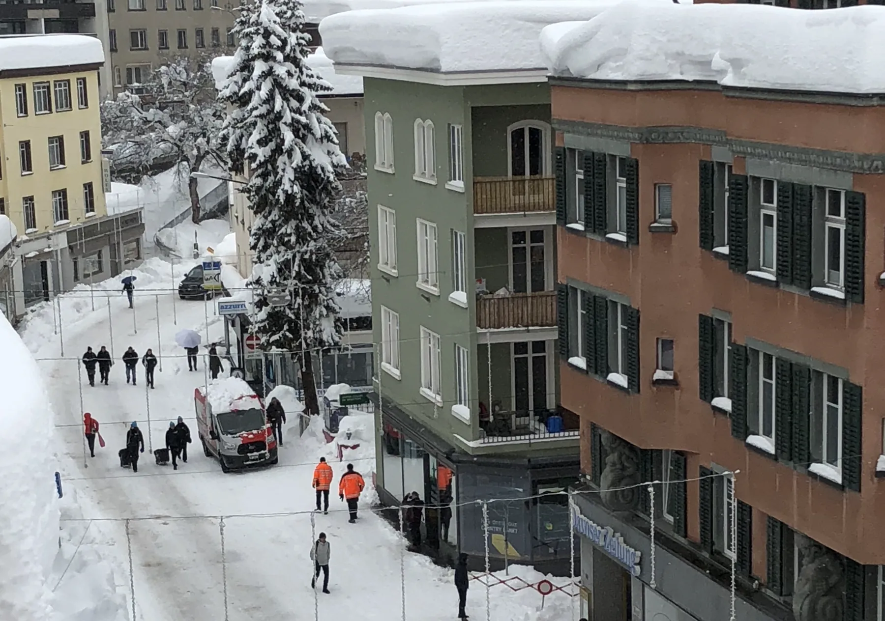 A group with wheeled luggage walking up a snow-lined street in Davos past security staff in orange jackets