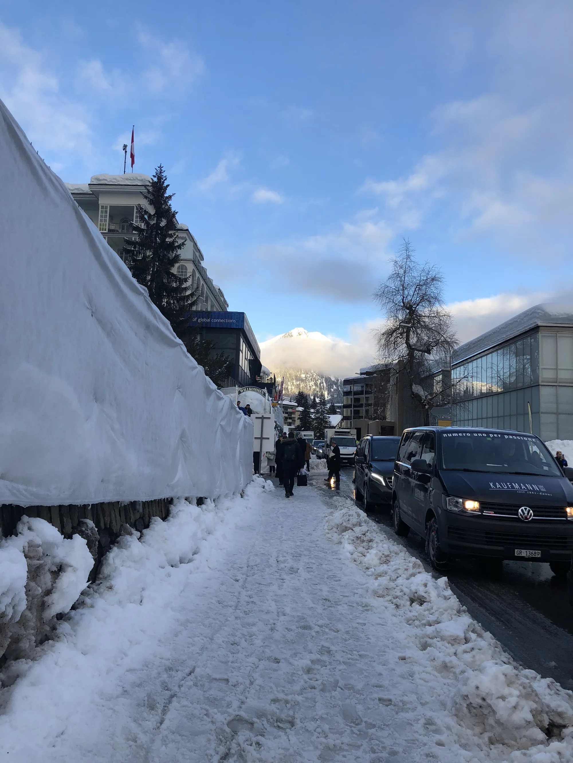 A snowy Davos street in daylight, cars parked along the roadside banked with cleared snow