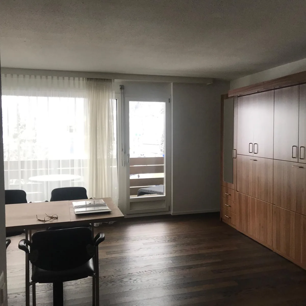 A studio with a meeting table and chairs by a balcony door, sheer curtains and a wall of wooden cabinets